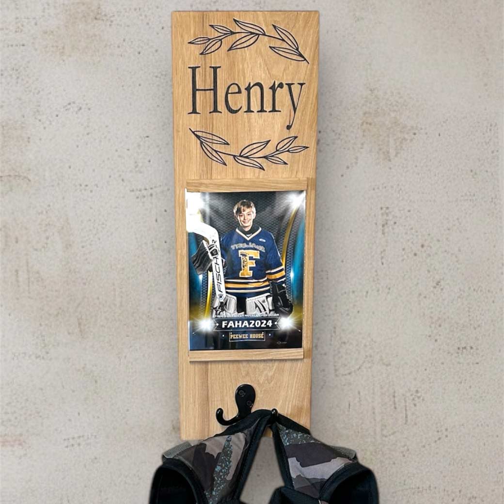 A personalized wooden coat and backpack rack with a name printed at the top, featuring a double hook for hanging items and a frame for a photo display.