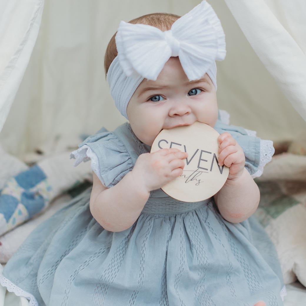 Baby in blue dress holding milestone cards from Birchmark Designs showing 7 months