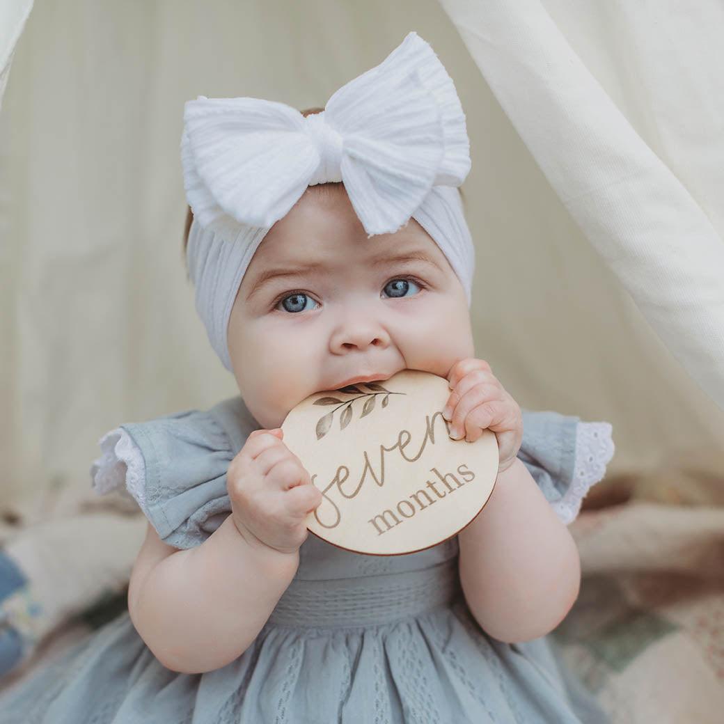 Baby in gray dress holding olive baby milestone card at 7 months